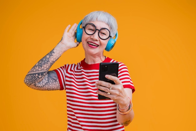 Cuffie e smartphone per musica d'ascolto femminile anziana anziana mentre si muovono danza emozione spensierata positiva isolata su sfondo giallo donna asiatica con tatuaggio musica felice danza canta studio shot