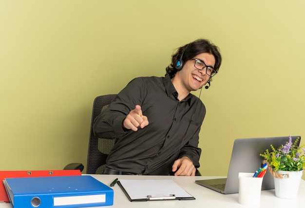 Cuffie manon di lavoratore di ufficio sorridente giovane in vetri ottici si siede alla scrivania con strumenti per ufficio utilizzando laptop che punta alla telecamera isolata su sfondo verde con spazio di copia