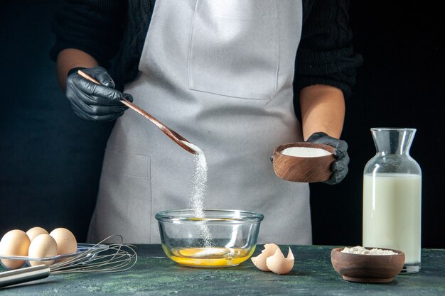 Cuoca di vista frontale che versa la farina nelle uova per l'impasto sul lavoro di cucina dell'operaio del forno della torta della torta della pasticceria scura