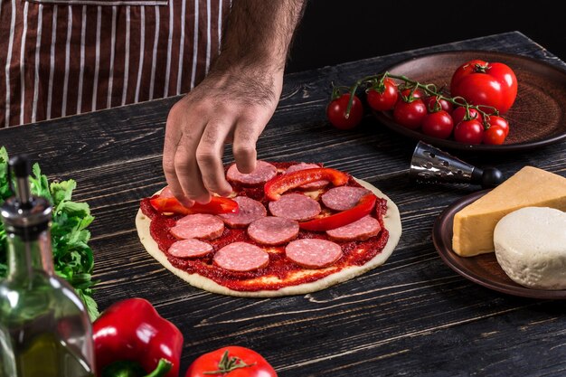 Cuocere in cucina mettendo gli ingredienti sulla pizza su un vecchio fondo di legno. Concetto di pizza. Produzione e consegna di cibo. Concetto di cucina. Avvicinamento