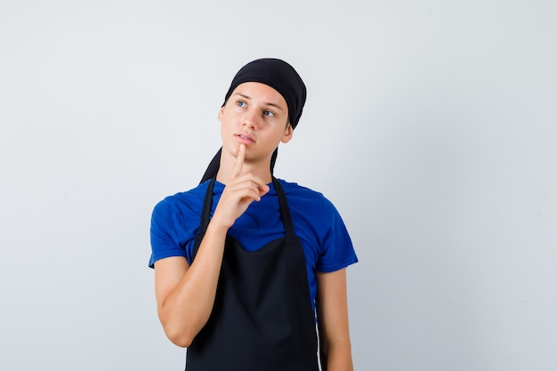 Cuoco adolescente maschio che tiene il dito sul mento in maglietta, grembiule e sembra preoccupato. vista frontale.