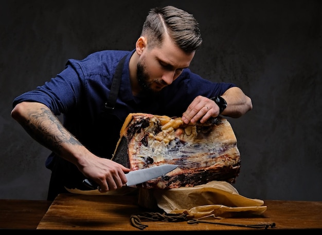 Cuoco concentrato che taglia carne a scatti esclusiva su un tavolo in una casa di caccia su uno sfondo scuro.