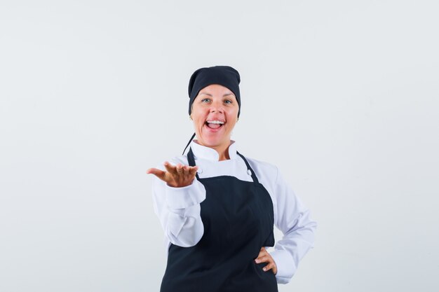 Cuoco femminile che allunga la mano come presentando qualcosa in uniforme, grembiule e guardando fiducioso, vista frontale.