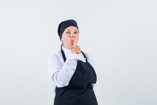 Cuoco femminile che mostra gesto di silenzio in uniforme, grembiule e guardando attento, vista frontale.