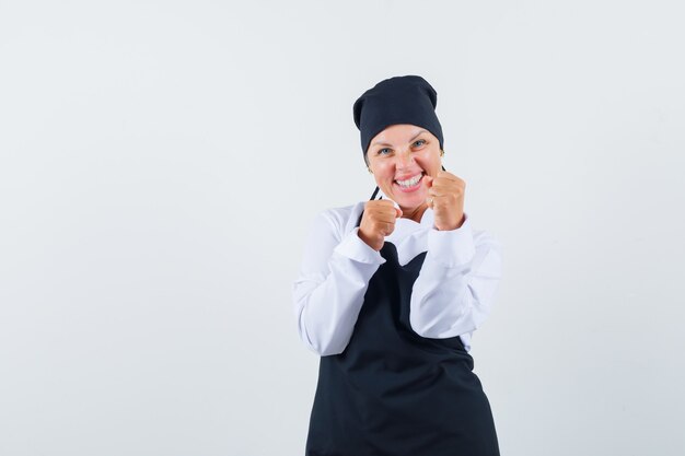 Cuoco femminile che sta nella posa di lotta in uniforme, grembiule e che sembra fortunato, vista frontale.