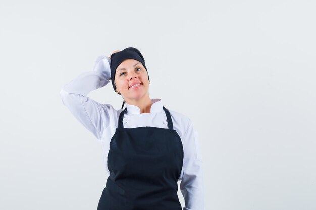 Cuoco femminile che tiene la mano dietro la testa in uniforme, grembiule e sembra sognante, vista frontale.