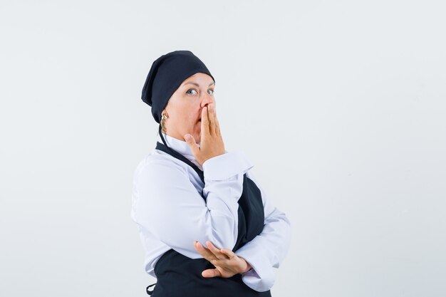 Cuoco femminile che tiene la mano sulla bocca in uniforme, grembiule e che sembra sorpreso. vista frontale.