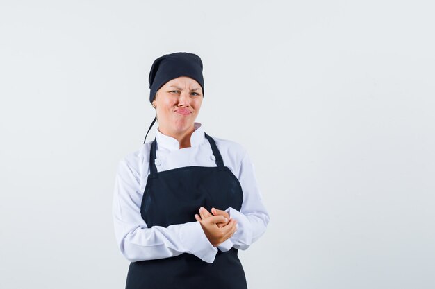 Cuoco femminile che tiene le mani giunte in uniforme, grembiule e che sembra insoddisfatto. vista frontale.