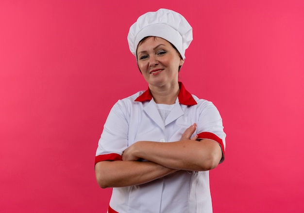 Cuoco femminile di mezza età sicuro nelle mani dell'incrocio dell'uniforme del cuoco unico sulla parete rosa isolata con lo spazio della copia