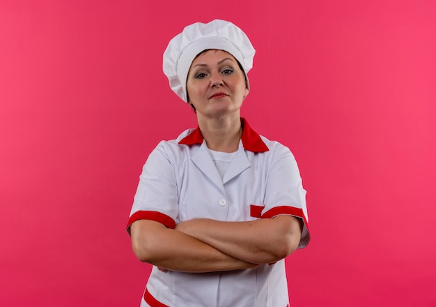 Cuoco femminile di mezza età sicuro nelle mani dell'incrocio dell'uniforme del cuoco unico sulla parete rosa isolata con lo spazio della copia