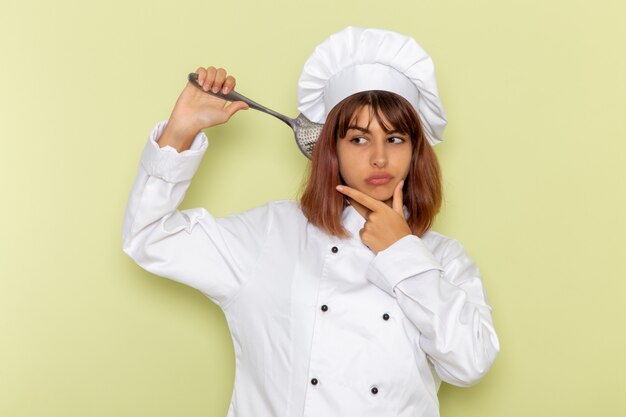 Cuoco femminile di vista frontale in vestito bianco del cuoco che tiene il grande cucchiaio d'argento sulla superficie verde chiaro