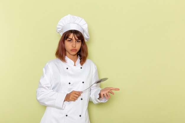 Cuoco femminile di vista frontale in vestito bianco del cuoco che tiene il grande cucchiaio d'argento sulla superficie verde