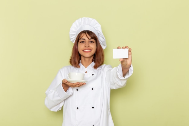 Cuoco femminile di vista frontale in vestito bianco del cuoco che tiene tazza di tè e carta sulla superficie verde