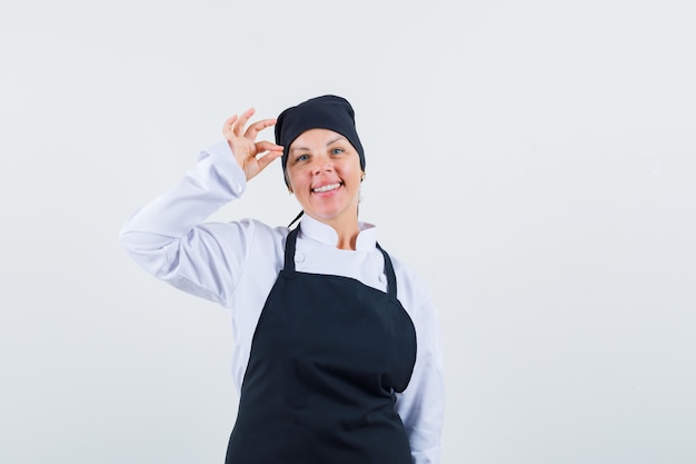 Cuoco femminile in uniforme, grembiule che mostra gesto giusto e che sembra allegro, vista frontale.