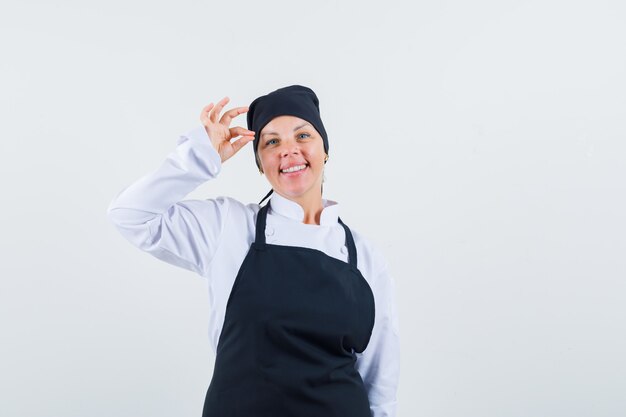 Cuoco femminile in uniforme, grembiule che mostra gesto giusto e che sembra allegro, vista frontale.