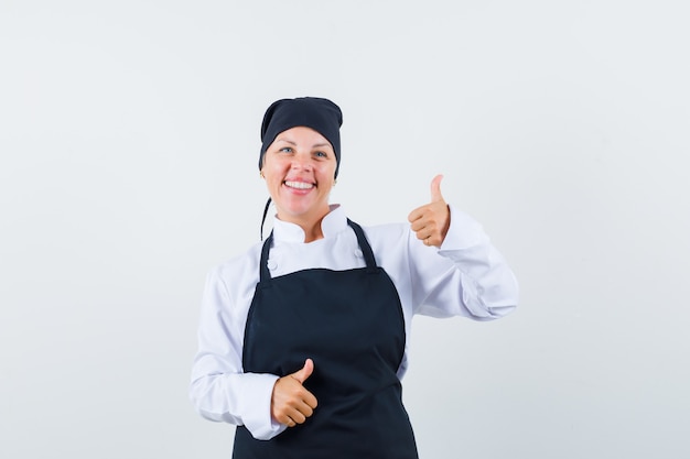 Cuoco femminile in uniforme, grembiule che mostra i doppi pollici in su e che sembra contento, vista frontale.