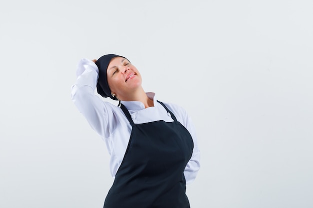 Cuoco femminile in uniforme, grembiule che tiene la mano sulla testa e che sembra felice, vista frontale.