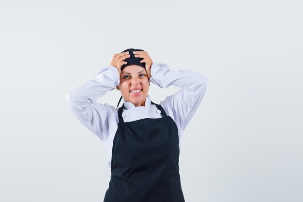 Cuoco femminile in uniforme, grembiule che tiene le mani sulla testa e che sembra smemorato, vista frontale.