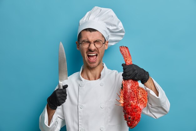 Cuoco francese professionista arrabbiato trascorre molto tempo in cucina, indossa uniformi, guanti di gomma neri, posa con pesce e coltello