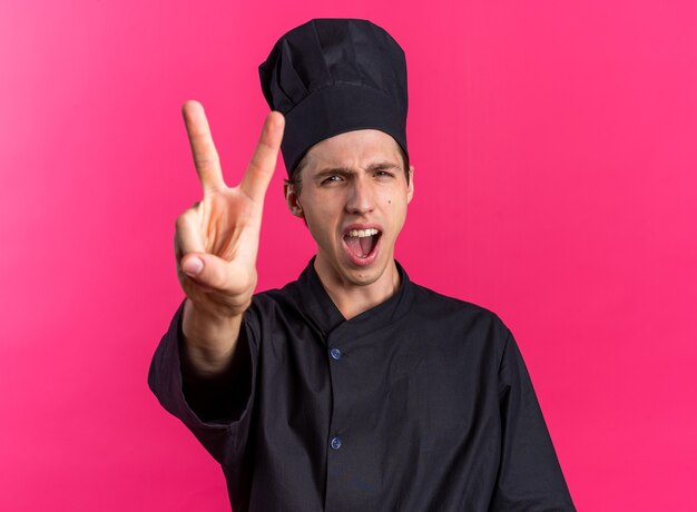 Cuoco maschio biondo rigoroso in uniforme da chef e berretto che guarda la telecamera urlando mostrando due con la mano isolata sulla parete rosa