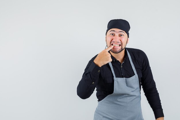 Cuoco maschio che indica ai suoi denti in camicia, grembiule, vista frontale.