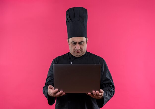 Cuoco maschio di mezza età confuso in uniforme del cuoco unico che esamina il computer portatile in sua mano sulla parete rosa isolata con lo spazio della copia