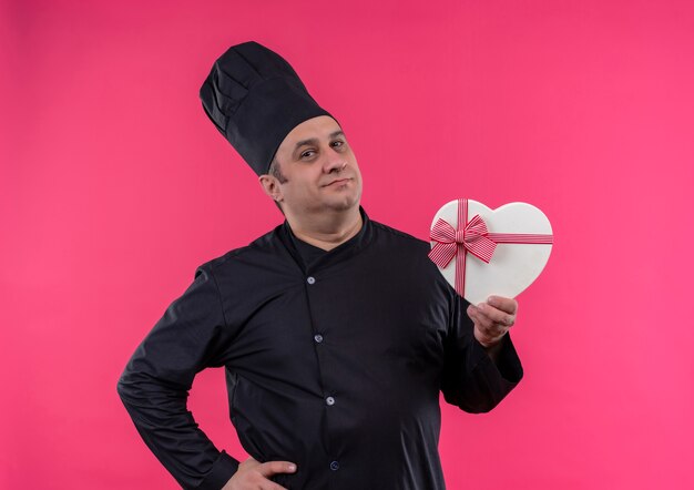 Cuoco maschio di mezza età in uniforme da chef tenendo la scatola a forma di cuore mise la mano sul fianco sul muro rosa isolato