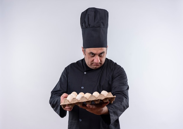 cuoco maschio di mezza età in uniforme del cuoco unico che tiene fuori lotto di uova sul muro bianco isolato