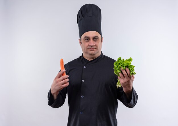 cuoco maschio di mezza età in uniforme del cuoco unico che tiene la carota e l'insalata sulla parete bianca isolata