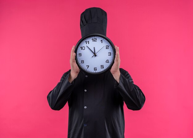 Cuoco maschio di mezza età in uniforme del cuoco unico coperto il fronte con l'orologio di parete sulla parete rosa isolata con lo spazio della copia
