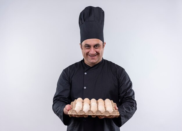 Cuoco maschio di mezza età sorridente in uniforme del cuoco unico che tiene fuori il lotto di uova alla macchina fotografica con lo spazio della copia