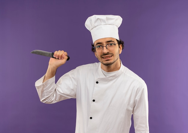 Cuoco maschio giovane teso che indossa l'uniforme del cuoco unico e vetri che tengono coltello sulla porpora