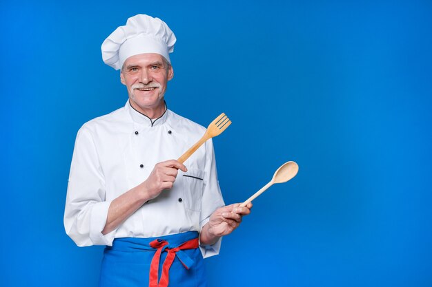 Cuoco unico anziano positivo che tiene cucchiaio e forchetta di legno, in uniforme bianca e cappuccio in posa isolata sulla parete blu
