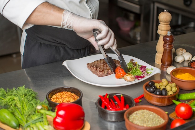 Cuoco unico che prepara la bistecca di manzo per il servizio con insalata e verdure grigliate