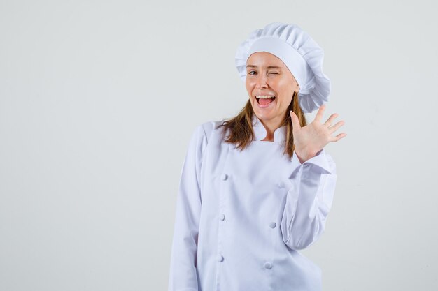 Cuoco unico femminile agitando la mano con gli occhi sbattuti in uniforme bianca e guardando energico