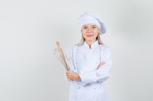 Cuoco unico femminile che tiene cucchiaio di legno, frusta, mattarello in uniforme bianca e che sembra allegro.