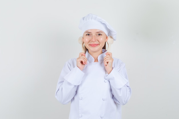 Cuoco unico femminile che tiene i cucchiai di legno dietro le orecchie in uniforme bianca e che sembra allegro