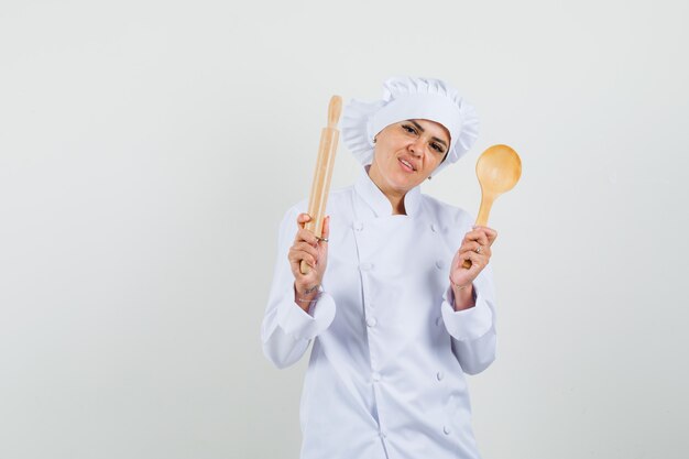 Cuoco unico femminile che tiene il mattarello e il cucchiaio di legno in uniforme bianca e che sembra fiducioso
