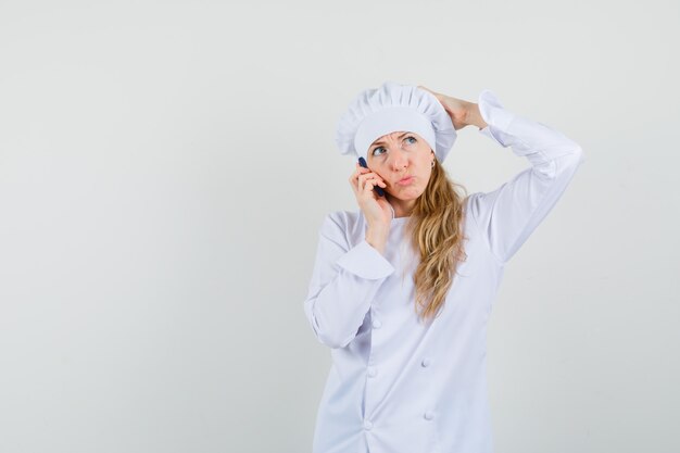 Cuoco unico femminile graffiare la testa mentre parla al cellulare in uniforme bianca e guardando pensieroso.