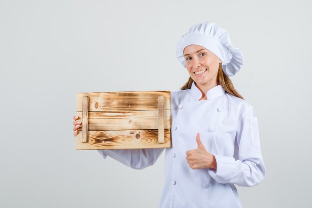 Cuoco unico femminile in uniforme bianca che tiene vassoio di legno con il pollice in su e che sembra allegro