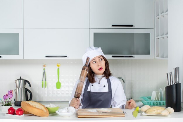 Cuoco unico femminile sorpreso in uniforme in piedi dietro il tavolo che prepara la pasticceria nella cucina bianca