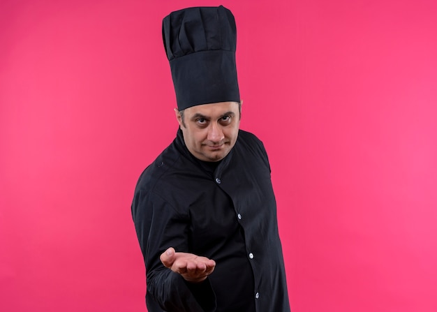 Cuoco unico maschio che indossa l'uniforme nera e cappello da cuoco che guarda l'obbiettivo che offre la mano greenting in piedi su sfondo rosa