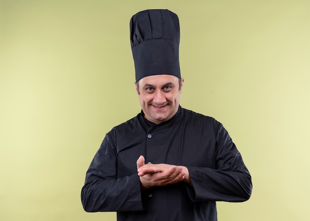 Cuoco unico maschio che indossa l'uniforme nera e cappello da cuoco che guarda l'obbiettivo con un sorriso sornione tenendo le mani insieme in piedi su sfondo verde