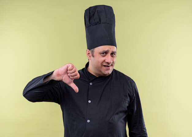 Cuoco unico maschio che indossa l'uniforme nera e cappello da cuoco che guarda l'obbiettivo scontento che mostra i pollici giù in piedi su sfondo verde