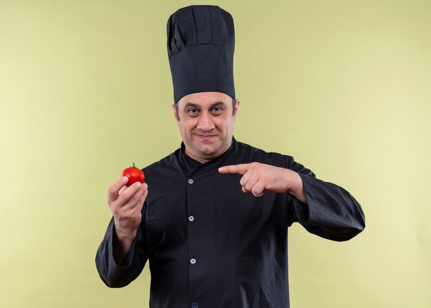 Cuoco unico maschio che indossa l'uniforme nera e cappello da cuoco che mostra il pomodoro che punta con il dito ad esso sorridendo guardando la fotocamera in piedi su sfondo verde