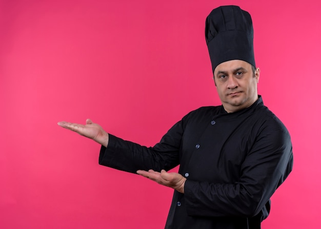 Cuoco unico maschio che indossa l'uniforme nera e cappello da cuoco che presenta qualcosa con le braccia in piedi su sfondo rosa