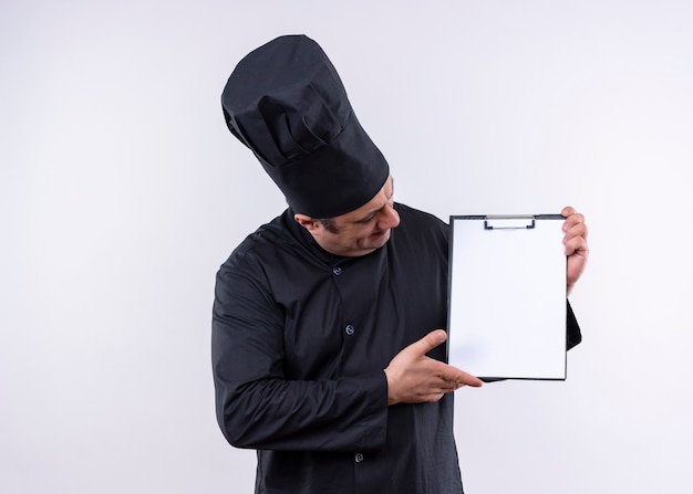 Cuoco unico maschio che indossa l'uniforme nera e cappello da cuoco che tiene appunti con pagine bianche guardandolo in piedi su sfondo bianco