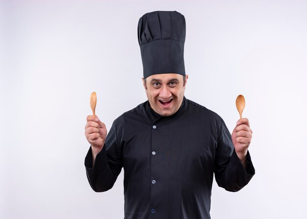 Cuoco unico maschio che indossa l'uniforme nera e cappello da cuoco che tiene due cucchiai di legno che guarda l'obbiettivo che sorride allegramente in piedi sopra priorità bassa bianca