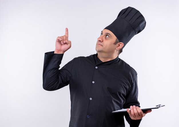 Cuoco unico maschio che indossa l'uniforme nera e cappello da cuoco che tiene gli appunti pointng con il dito in piedi su sfondo bianco