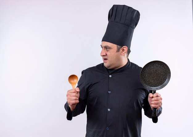 Cuoco unico maschio che indossa l'uniforme nera e cappello da cuoco che tiene il cucchiaio di legno e la padella che guarda da parte sorpreso in piedi su sfondo bianco